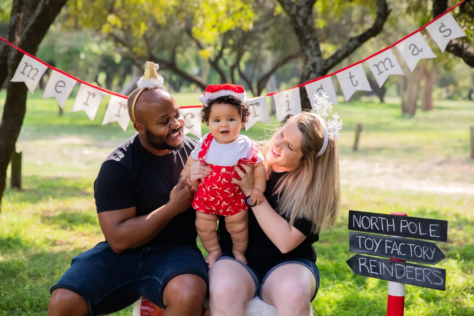 Parents holding up their baby in a Santa hat beside a North Pole signpost